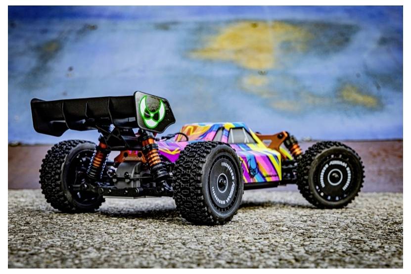 Ein buntes Spielzeugauto mit Spoiler und groben Profil-Reifen steht auf einer strukturierten Oberfläche. Der Hintergrund ist eine verschwommene blau-beige Wand.