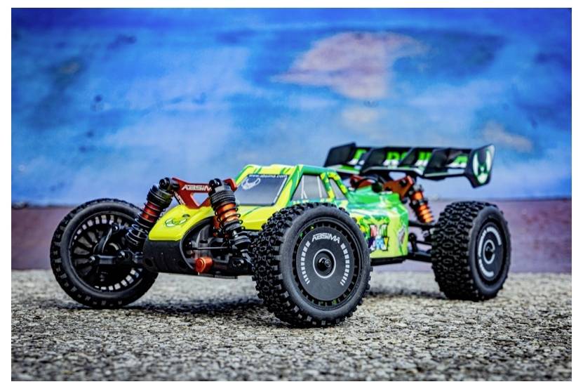 Ein ferngesteuertes Auto mit einem grün-gelben Karosseriebody, schwarzen Reifen und sichtbarer Federung, das auf einer Kiesoberfläche mit blauem Hintergrund steht.