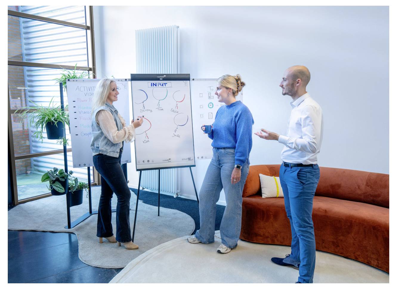 Drei Personen stehen um ein Flipchart herum und diskutieren über ein Diagramm. Die Büroumgebung umfasst ein Sofa und Pflanzen. Sie scheinen sich angeregt zu unterhalten.