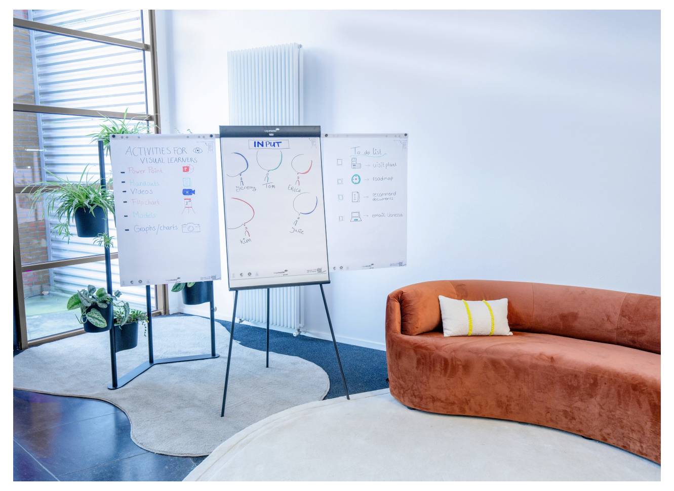 Ein moderner Besprechungsraum mit einem gebogenen orangefarbenen Sofa, zwei Flipcharts mit Diagrammen und Listen sowie einem großen Fenster im Hintergrund.