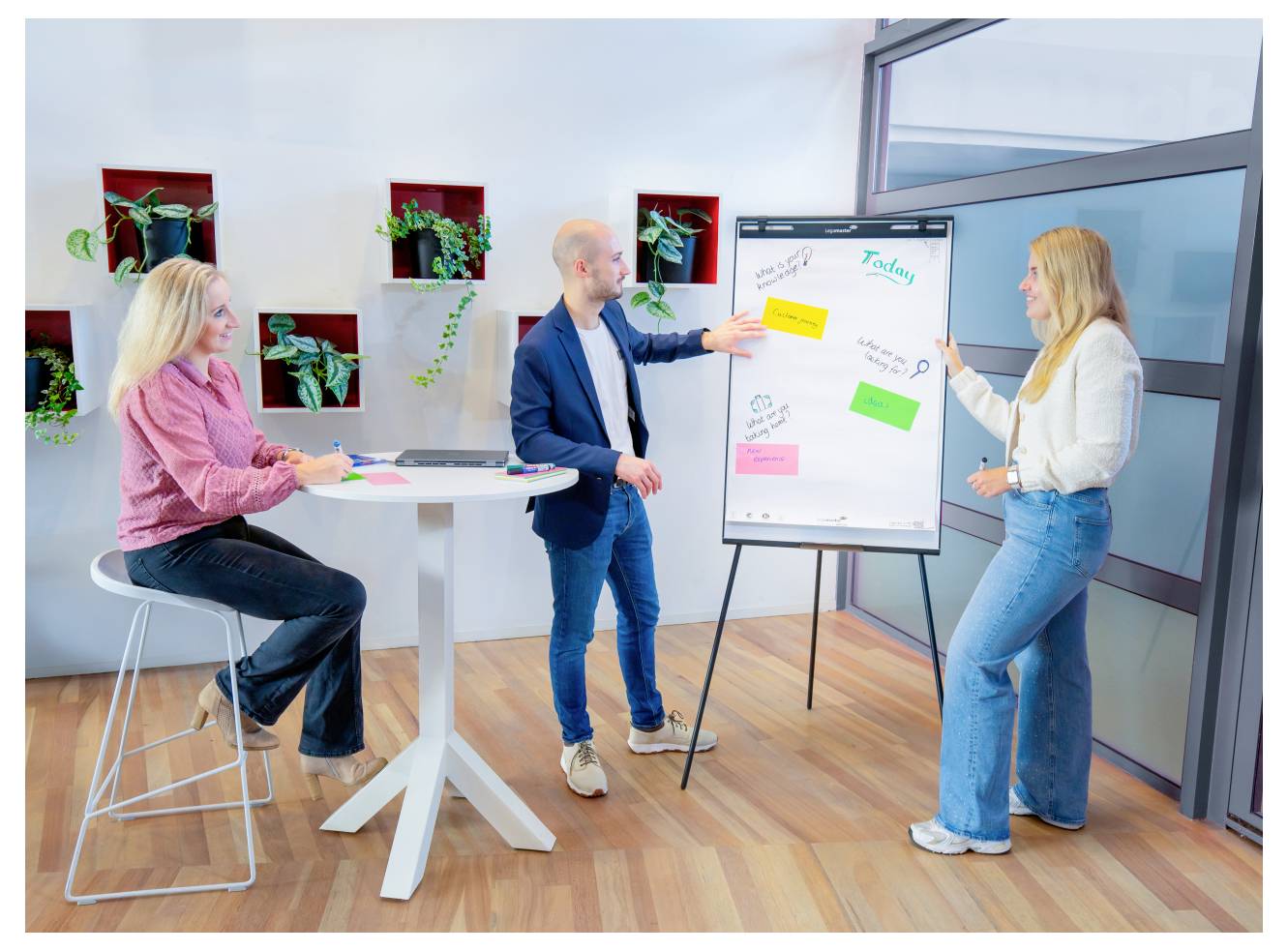 Drei Personen diskutieren Ideen in einem Büro. Eine Person zeigt auf ein Whiteboard mit Haftnotizen. Die anderen hören aufmerksam zu.