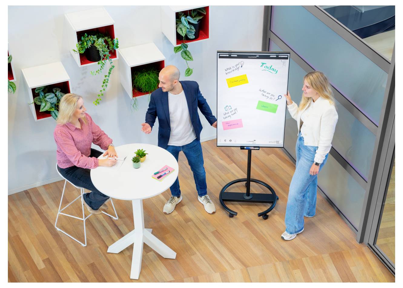Drei Personen diskutieren Ideen in einem Büroraum, wobei zwei neben einem Flipchart stehen und eine Person an einem runden Tisch sitzt.