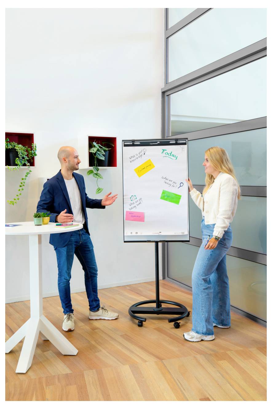 Ein Mann und eine Frau diskutieren Ideen in einem modernen Büro und zeigen auf bunte Haftnotizen auf einem großen Whiteboard an einem Rollständer.