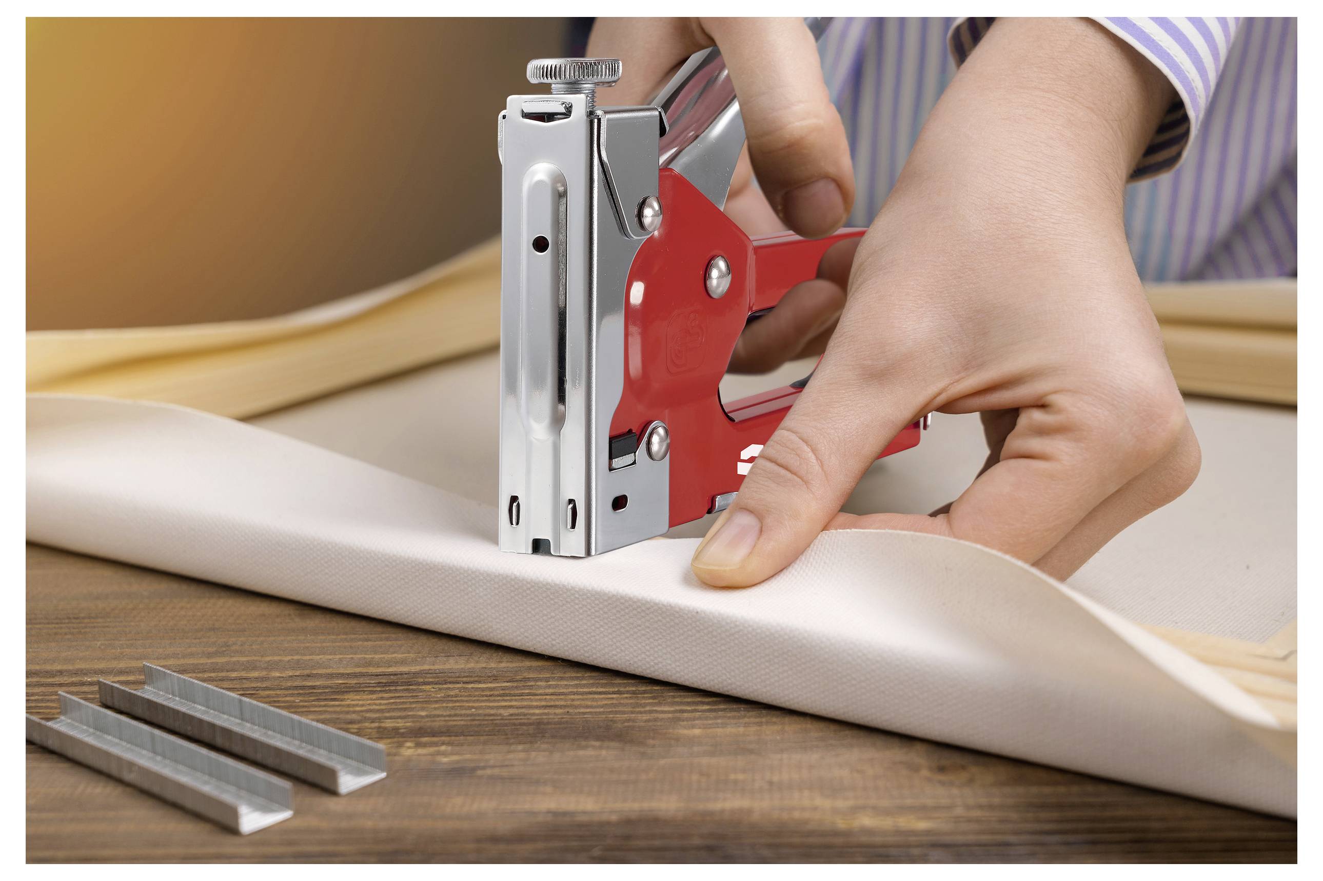 „Eine Person benutzt eine rote Tacker-Pistole, um Leinwand an einem Holzrahmen auf einem Tisch zu befestigen. Metallklammern liegen in der Nähe.