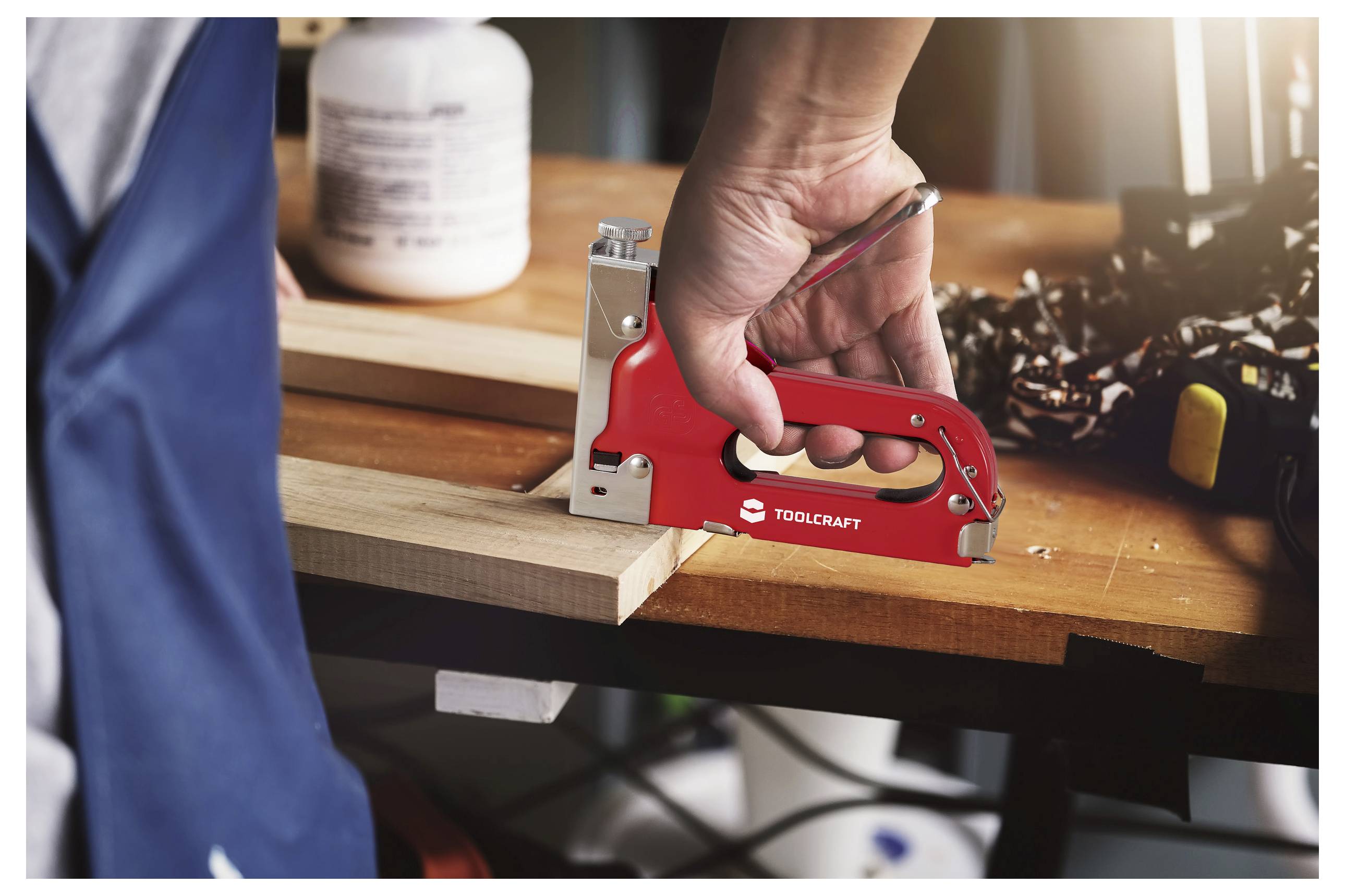 Eine Person, die eine rote Heftmaschine verwendet, um Holzbretter auf einer Werkbank zu befestigen, mit Werkzeugmaschinen und Ausrüstung im Hintergrund.