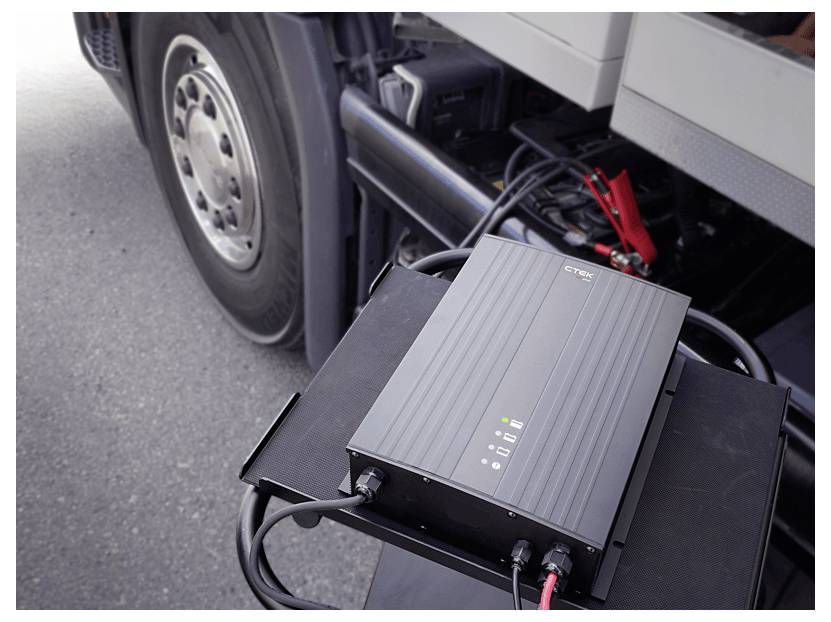 Ein schwarzes Ladegerät mit Kabeln, die an ein großes Fahrzeug angeschlossen sind. Das Rad des Fahrzeugs und eine Batterieklammer sind im Hintergrund sichtbar.