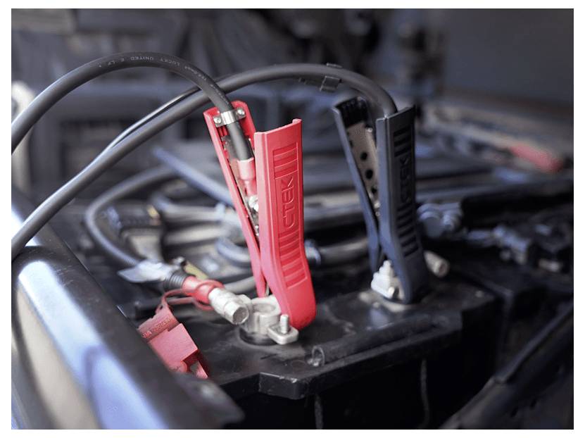 Eine Nahaufnahme einer Autobatterie mit roten und schwarzen Überbrückungskabeln, die einen Starthilfeprozess oder eine Batterieladung anzeigen.