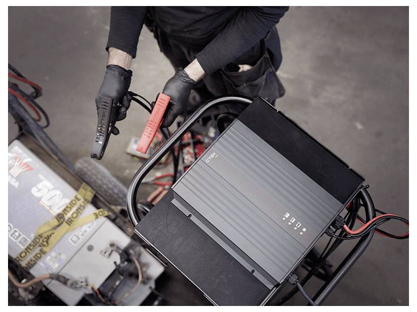 Eine Person mit Handschuhen, die Überbrückungskabel hält und sich darauf vorbereitet, ein Batterieladegerät an einem Fahrzeug in einer Garagenumgebung anzuschließen.