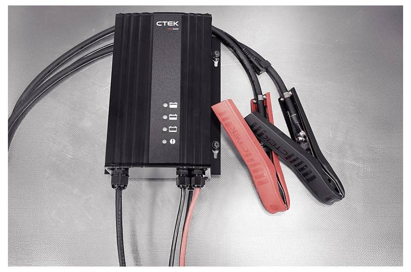 Ladegerät mit schwarzen und roten Klemmen auf einer metallischen Oberfläche. Das Ladegerät verfügt über vier Anzeigelichter und mehrere angeschlossene Kabel.