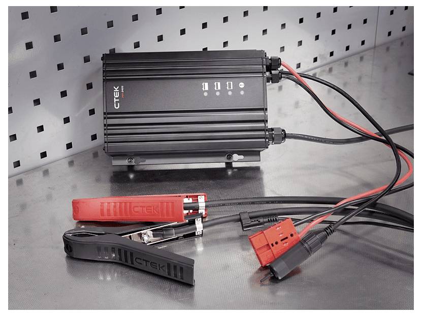 Ein Batterieladegerät mit der Bezeichnung 'CTEK MULTI US 25000' mit roten und schwarzen Kabeln und Klemmen, das auf einer Metalloberfläche neben einer Lochplatte liegt.