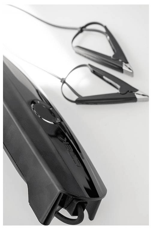 Ein schwarzer Starthilfe-Akku mit angebrachten Krokodilklemmen liegt diagonal auf einer weißen Oberfläche. Das Gerät hat ein schlankes Design.