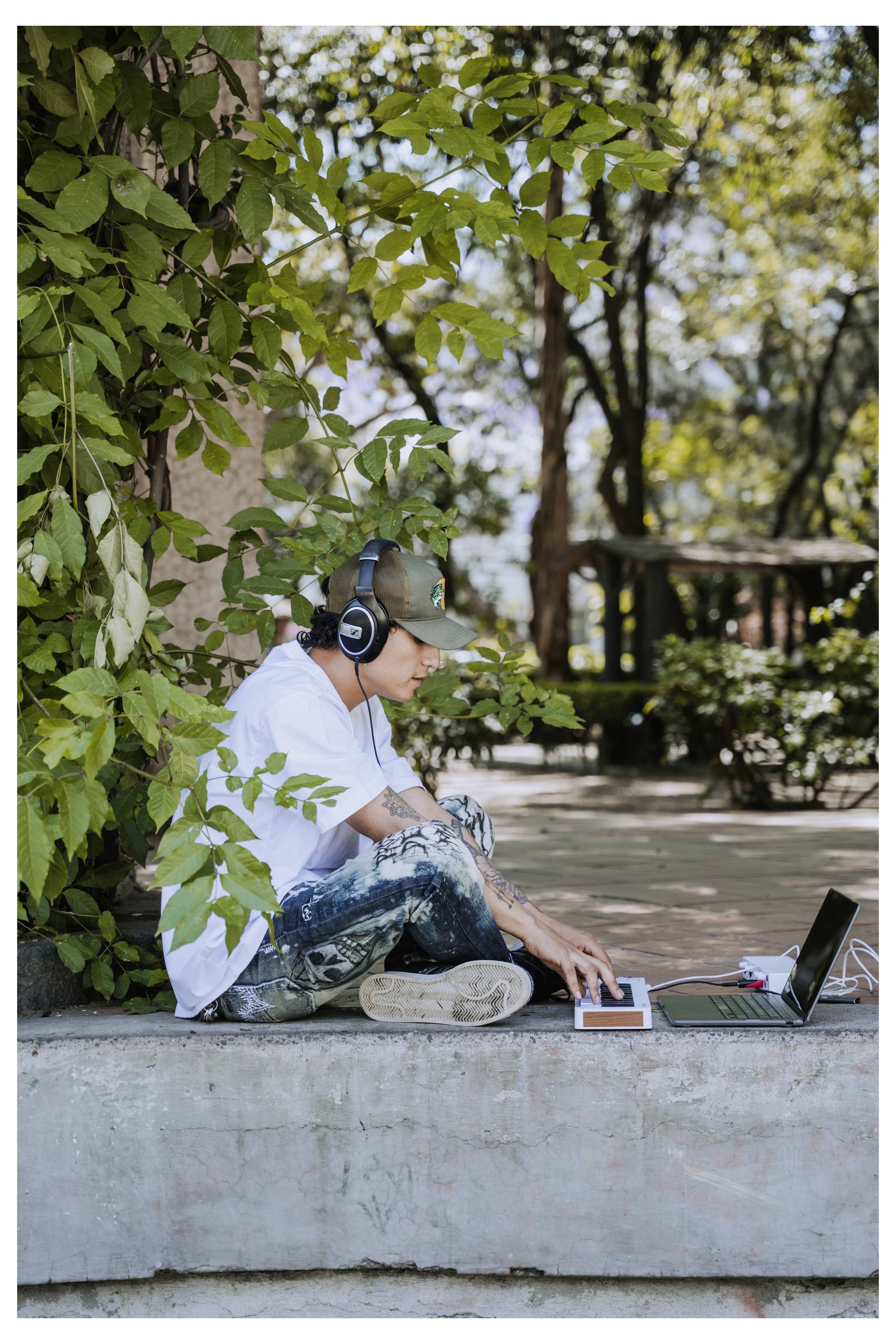 Eine Person mit Kopfhörern sitzt draußen auf einer Betonkante, arbeitet mit einem Laptop und einem Tablet, umgeben von Bäumen und Grünpflanzen.