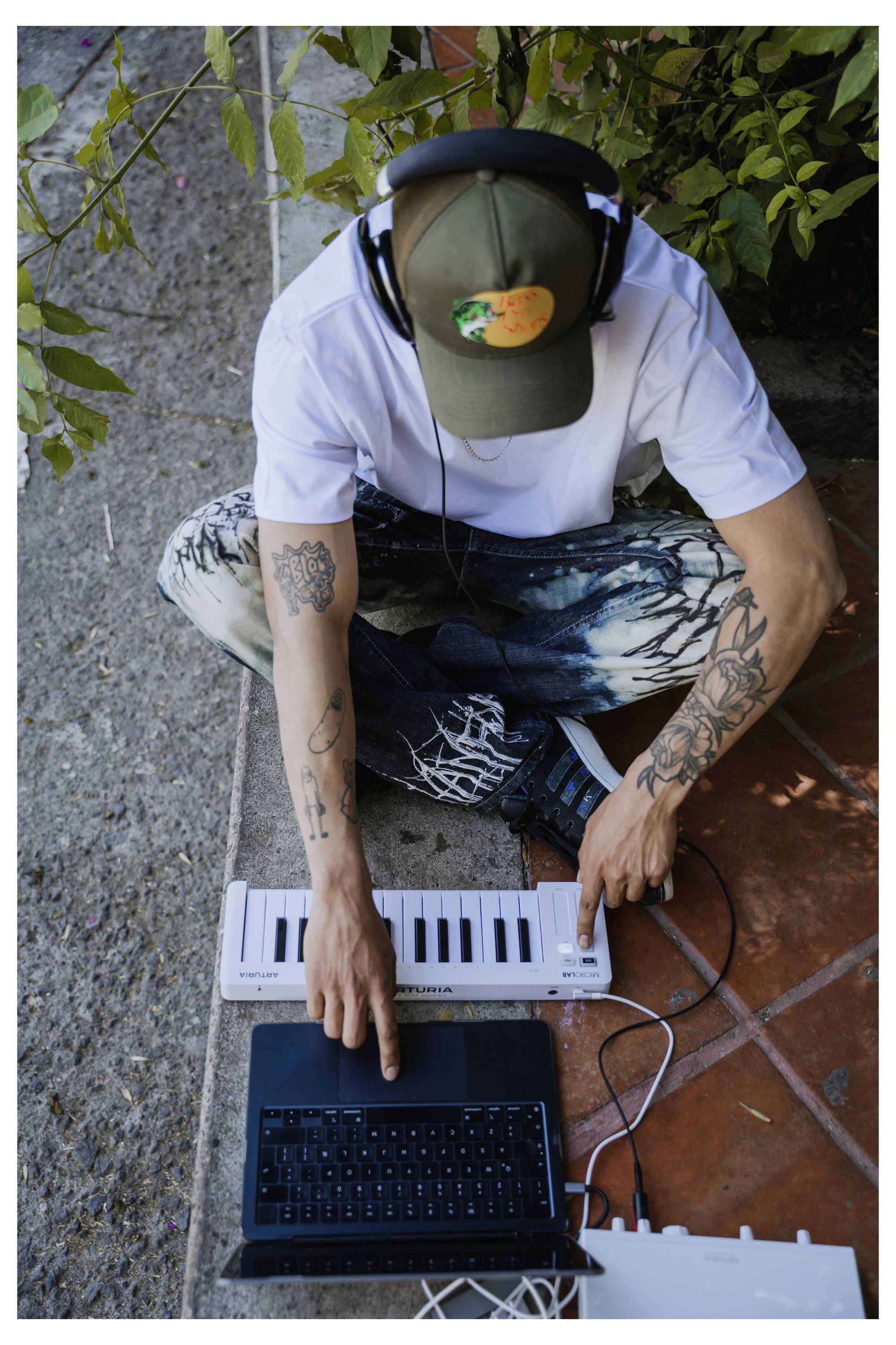 Eine Person mit Tattoos sitzt auf dem Boden, trägt Kopfhörer und eine Kappe und benutzt eine kleine Tastatur, die mit einem Laptop im Freien verbunden ist.