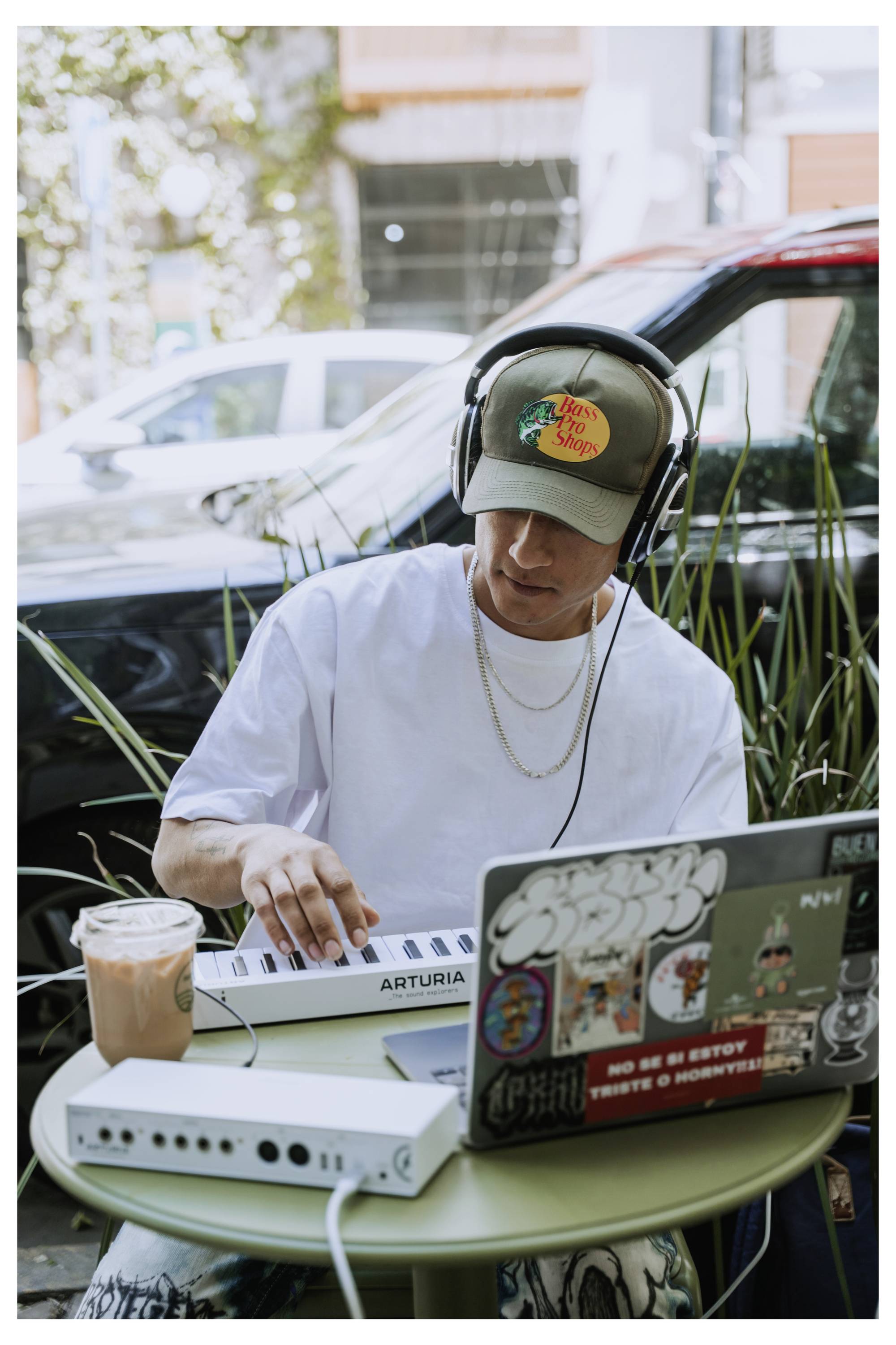 Eine Person mit Kopfhörern bedient einen Musikcontroller mit einem Laptop in einem Straßencafé. Ein Eiskaffee steht auf dem Tisch.