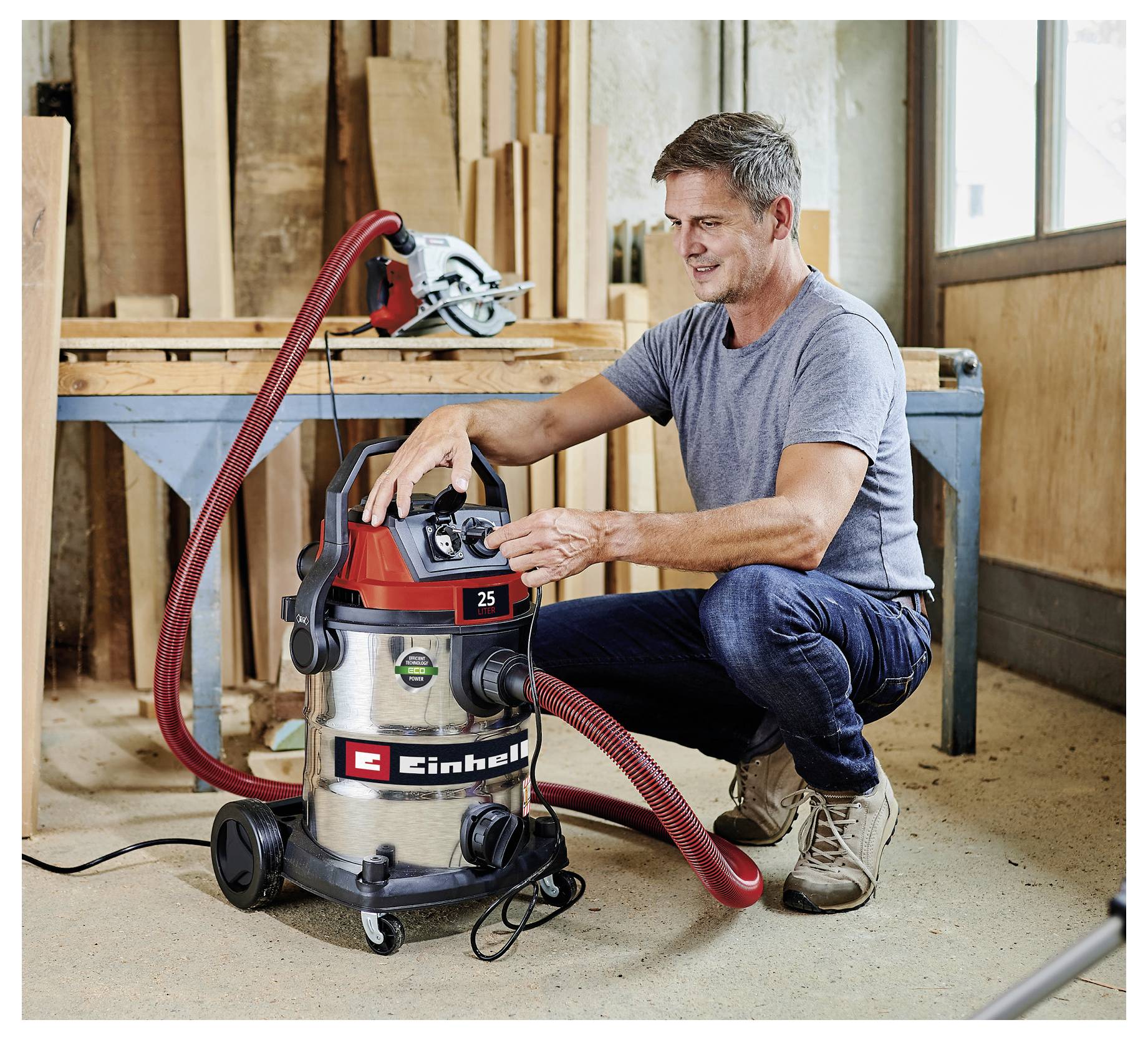 Eine Person bedient die Steuerungen eines Staubsaugers in einer Holzwerkstatt, umgeben von Werkzeugen und Holzbrettern, was eine aktive Vorbereitung zur Reinigung andeutet.