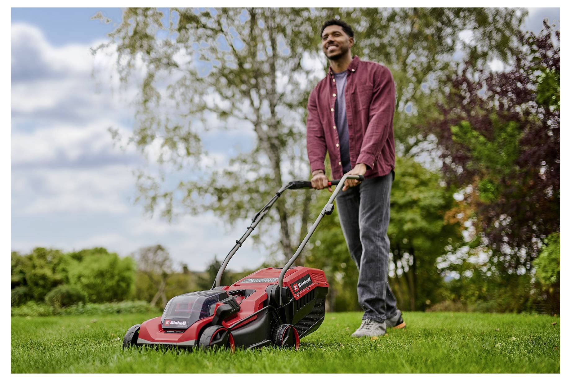Eine Person, die an einem teilweise bewölkten Tag mit einem roten elektrischen Rasenmäher in einem Garten Rasen mäht, lächelt und trägt lässige Kleidung.