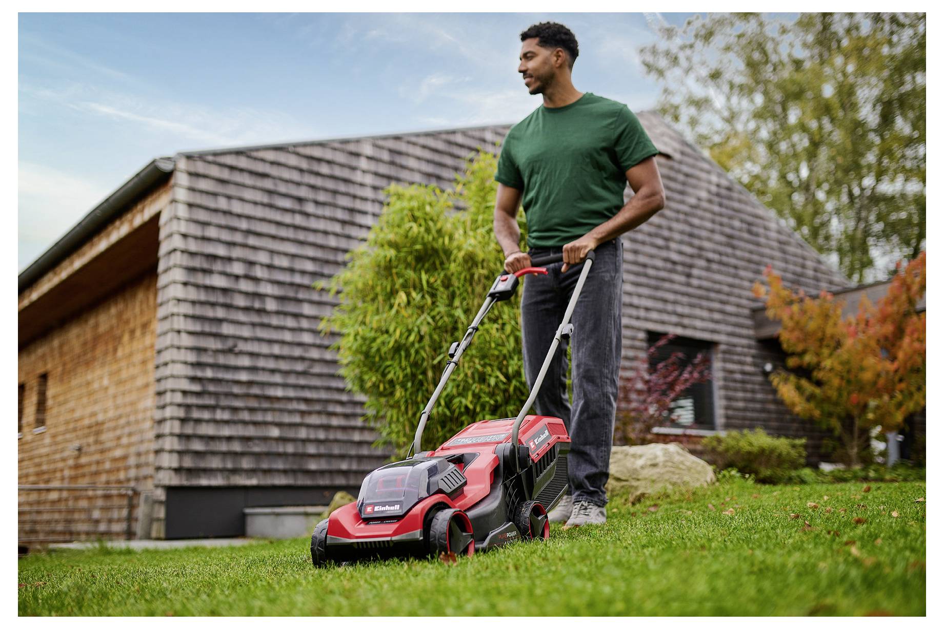 Eine Person in einem grünen Hemd schiebt einen roten Rasenmäher auf einer Rasenfläche vor einem Holzhaus, umgeben von Bäumen.