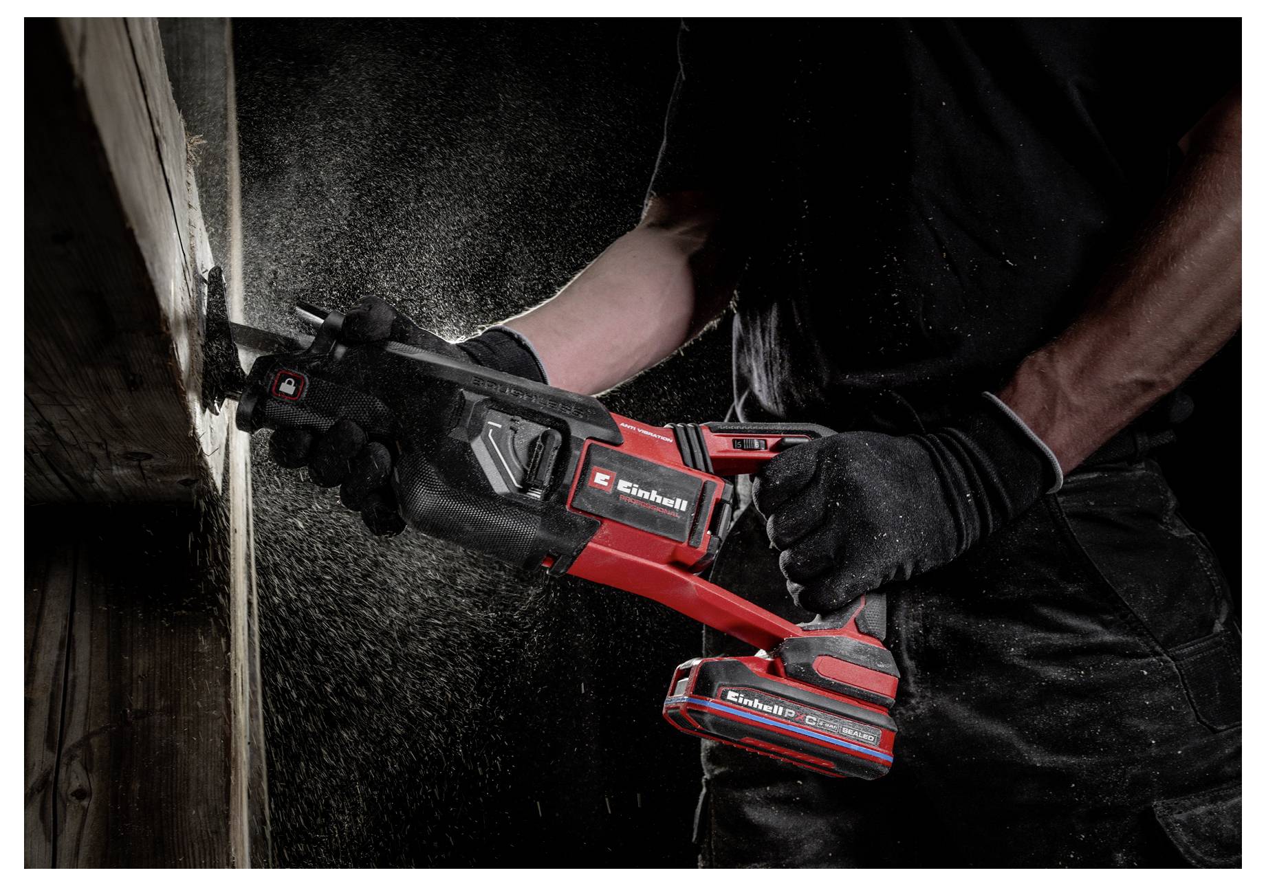 Eine Person mit Handschuhen benutzt eine rote und schwarze kabellose Säge, um einen Holzbalken durchzuschneiden, während Sägespäne durch die Luft fliegen.