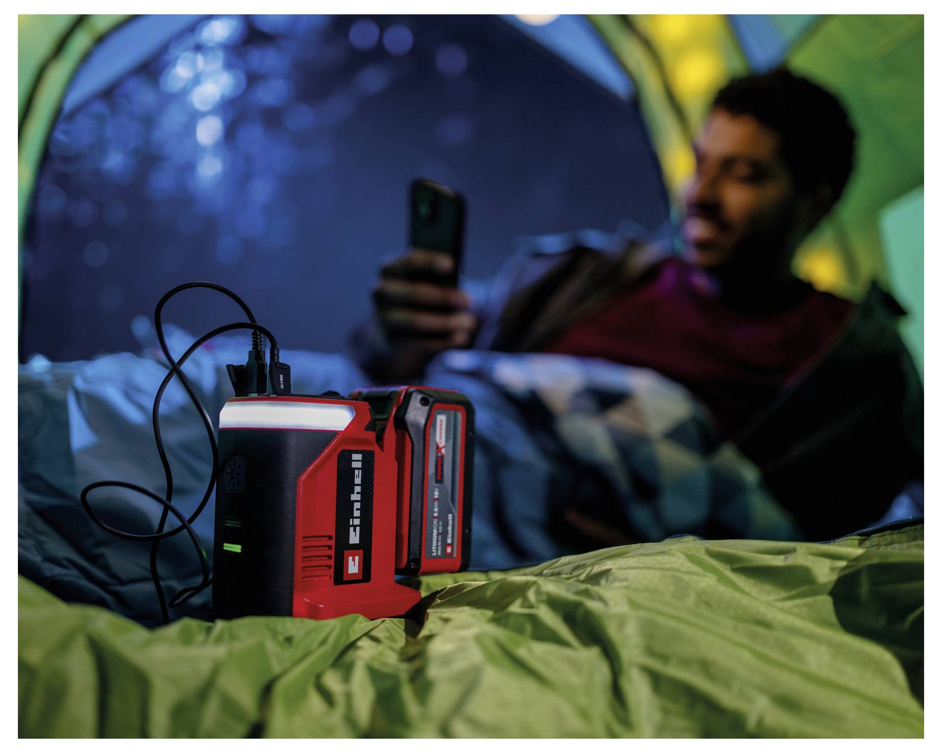 Eine Person in einem Zelt nutzt ein Smartphone, das mit einer roten mobilen Powerbank verbunden ist und damit den Einsatz von Camping-Technologie veranschaulicht.