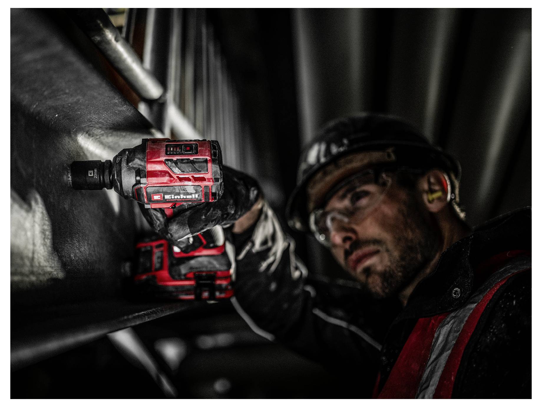 Ein Mann mit Sicherheitsausrüstung verwendet eine rote Bohrmaschine an einem Metallträger und betont damit Bau- oder industrielle Arbeit.