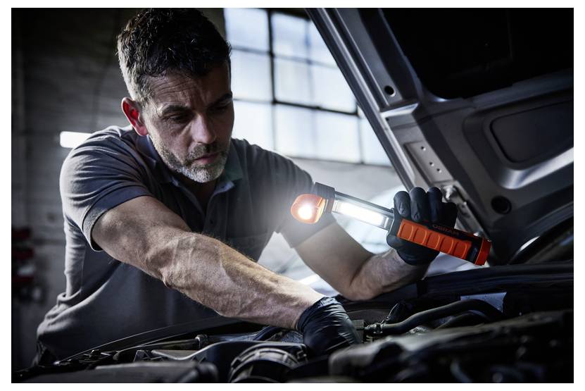 Ein Mechaniker untersucht einen Motor mit einer Taschenlampe in einer düster beleuchteten Garage, konzentriert auf Reparaturen, wobei die Motorhaube geöffnet über ihm ist.