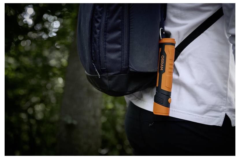 Eine Person mit einem schwarzen Rucksack trägt eine orangefarbene Taschenlampe, die daran befestigt ist, während sie durch einen Wald geht.
