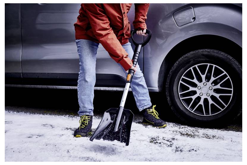 Eine Person mit einer roten Jacke und Jeans benutzt eine Schaufel, um Schnee von einer Einfahrt neben einem silbernen Auto zu räumen und konzentriert sich dabei auf die Winterreinigung.