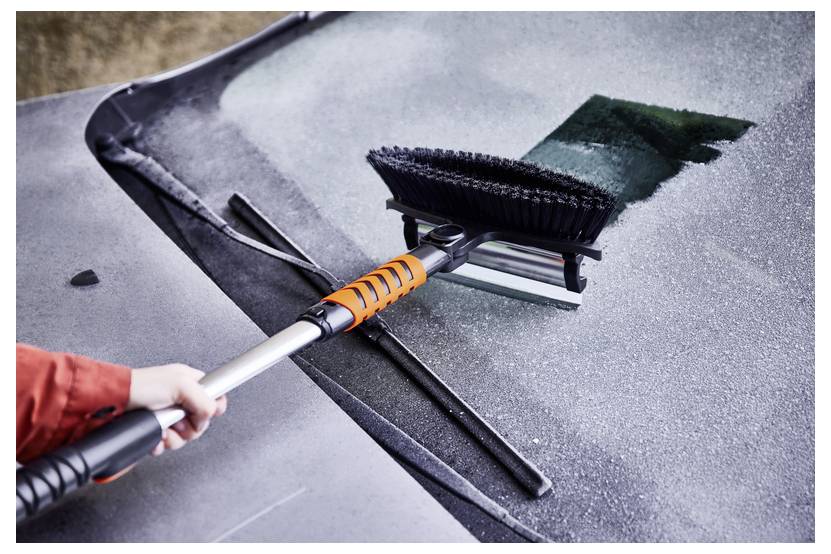 Eine Hand verwendet einen Abzieher mit einer Bürste, um Frost von der Windschutzscheibe eines Autos zu entfernen und dabei eine klare Stelle auf der eisigen Oberfläche freizulegen.