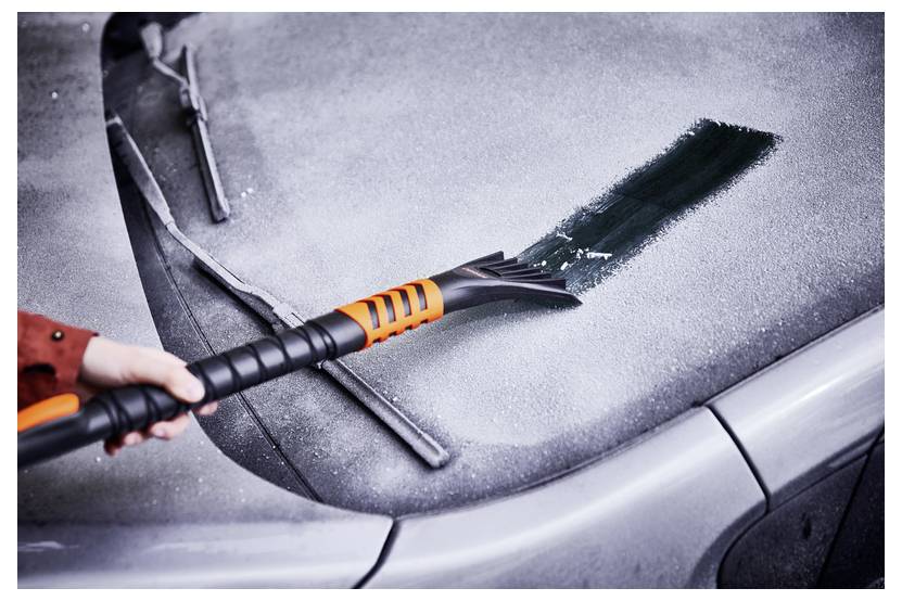 Eine Hand benutzt einen orangefarbenen Eiskratzer, um Frost von einer Autoscheibe zu entfernen und dabei einen sauberen Streifen an einem kalten, frostigen Morgen freizulegen.