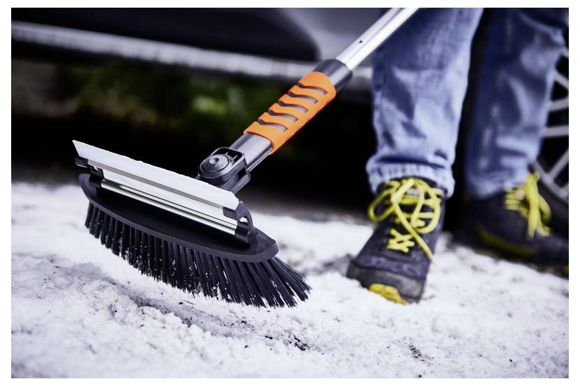 Eine Person fegt Schnee vom Boden mit einem Werkzeug, das einen Besen und einen Abzieher hat. Der Abzieher ist orange, und die Person trägt Schuhe mit gelben Schnürsenkeln.