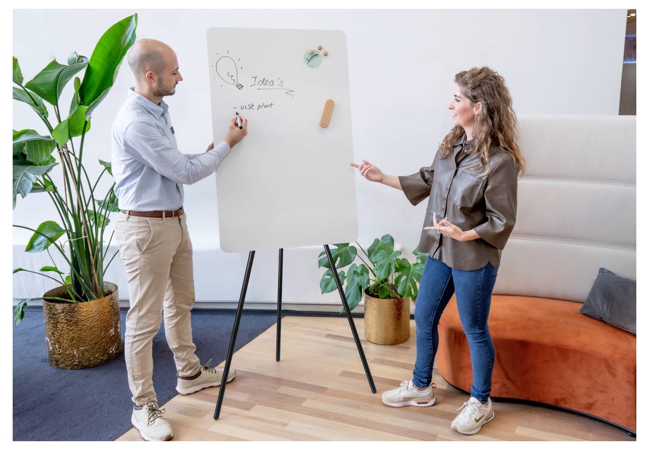 Eine Person präsentiert Ideen auf einem Whiteboard für eine andere Person in einer Büroumgebung, wobei im Hintergrund Zimmerpflanzen stehen.