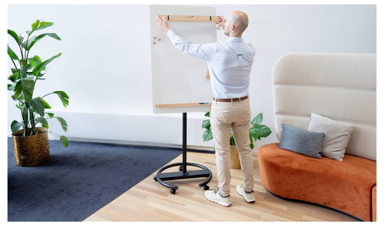 Eine Person steht in einem modernen Büroumfeld und schreibt auf ein Flipchart. Ein Sofa, Pflanzen und ein dunkler Teppich sind zu sehen.