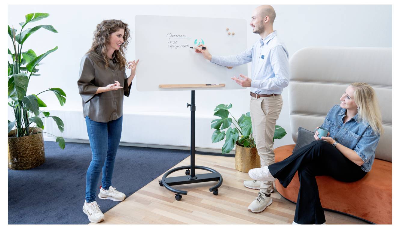 Drei Personen in einem gemütlichen Besprechungsraum; eine schreibt auf einem Whiteboard, während eine andere steht und gestikuliert, und die dritte Person sitzt und beobachtet.