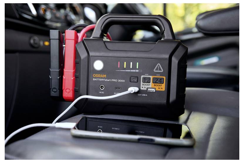 Ein tragbarer Starthilfe-Akku mit angeschlossenen Kabeln, der auf einem Autositz liegt. Ein Smartphone wird über einen USB-Anschluss am Gerät aufgeladen.