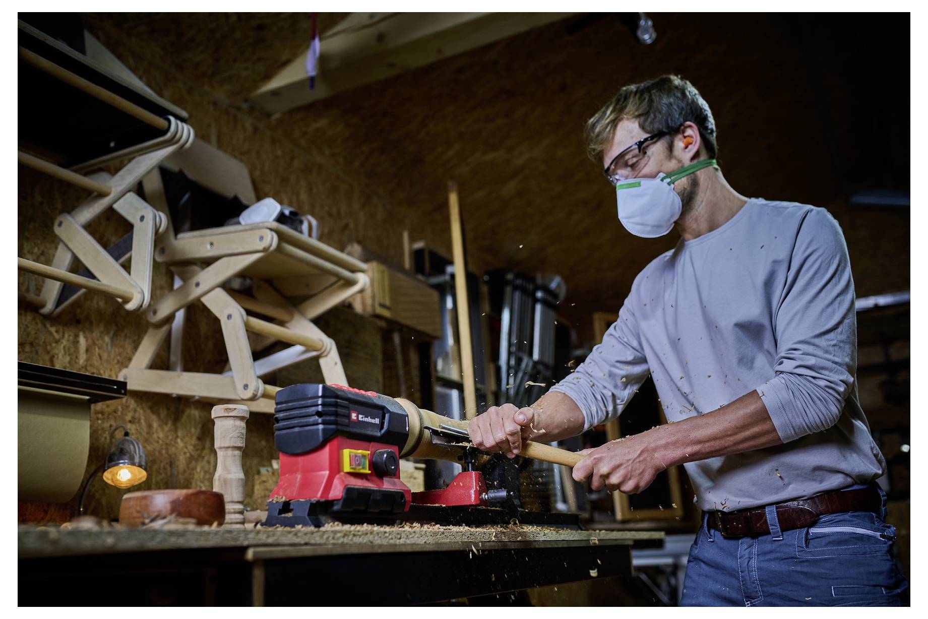 Eine Person mit Maske bearbeitet in einer Werkstatt ein Holzstück an einer Drechselbank. Im Hintergrund sind Werkzeuge und hölzerne Strukturen zu sehen.
