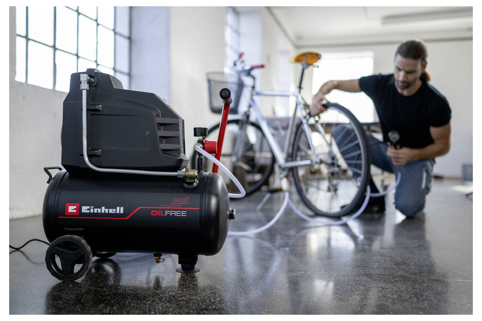 Ein Mann verwendet einen Luftkompressor, um einen Fahrradreifen in einem hellen Raum mit großen Fenstern aufzupumpen. Der Kompressor ist als 
