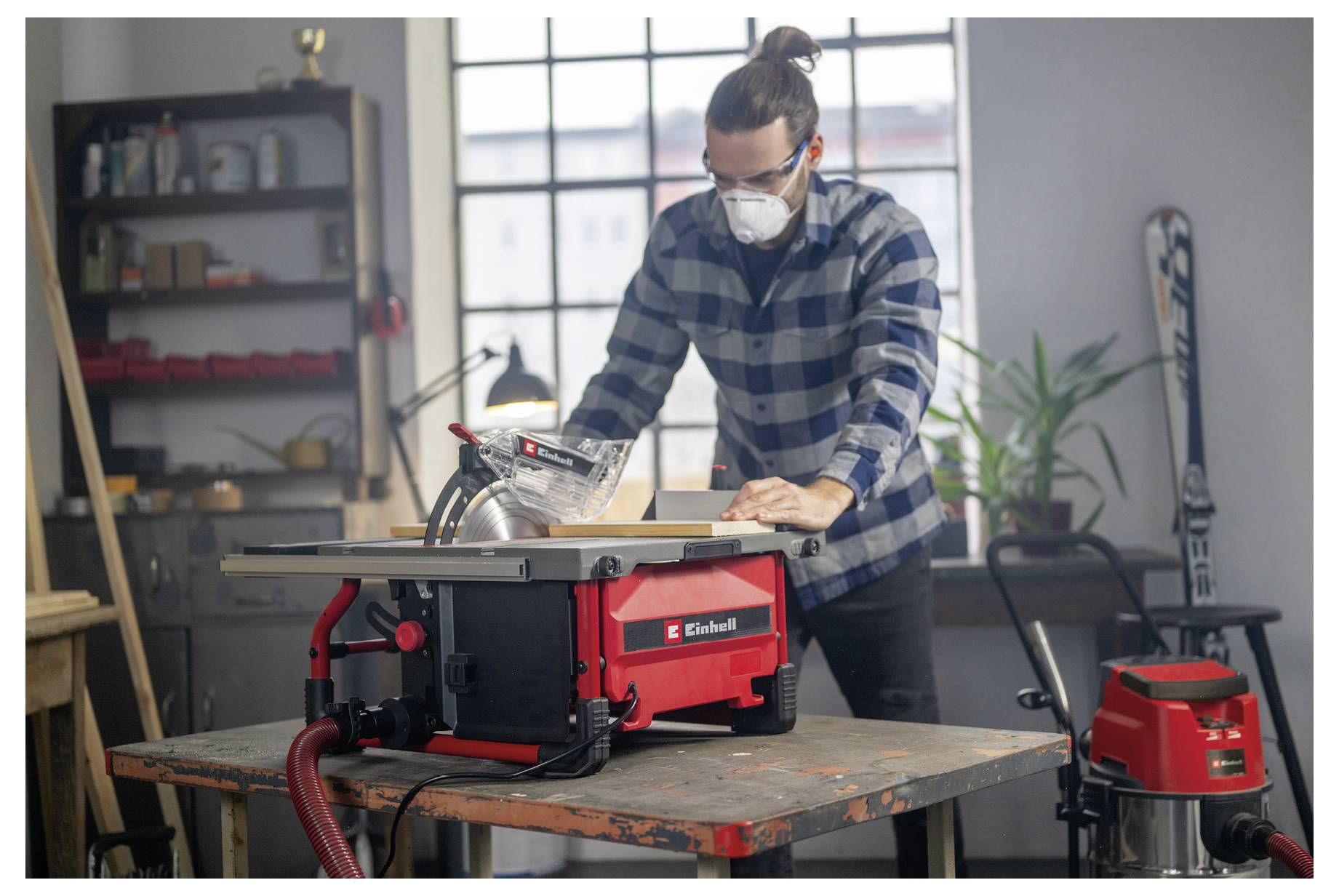 Eine Person mit Sicherheitsschutzbrille und kariertem Hemd bedient eine rote Tischkreissäge in einer Werkstatt, mit Regalen und Pflanzen im Hintergrund.