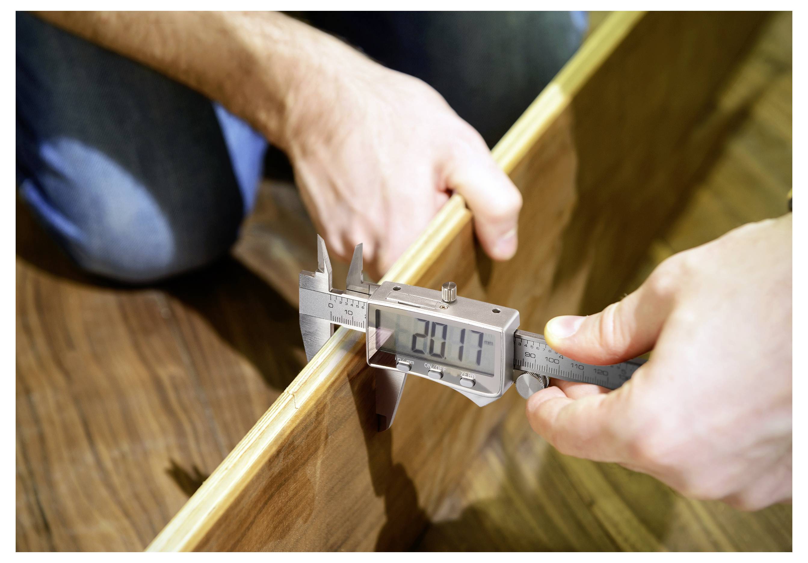 Eine Person misst die Dicke einer Holzplatte mit einer digitalen Schieblehre, die '20,17 mm' anzeigt, auf einem Holzoberflächen-Hintergrund.