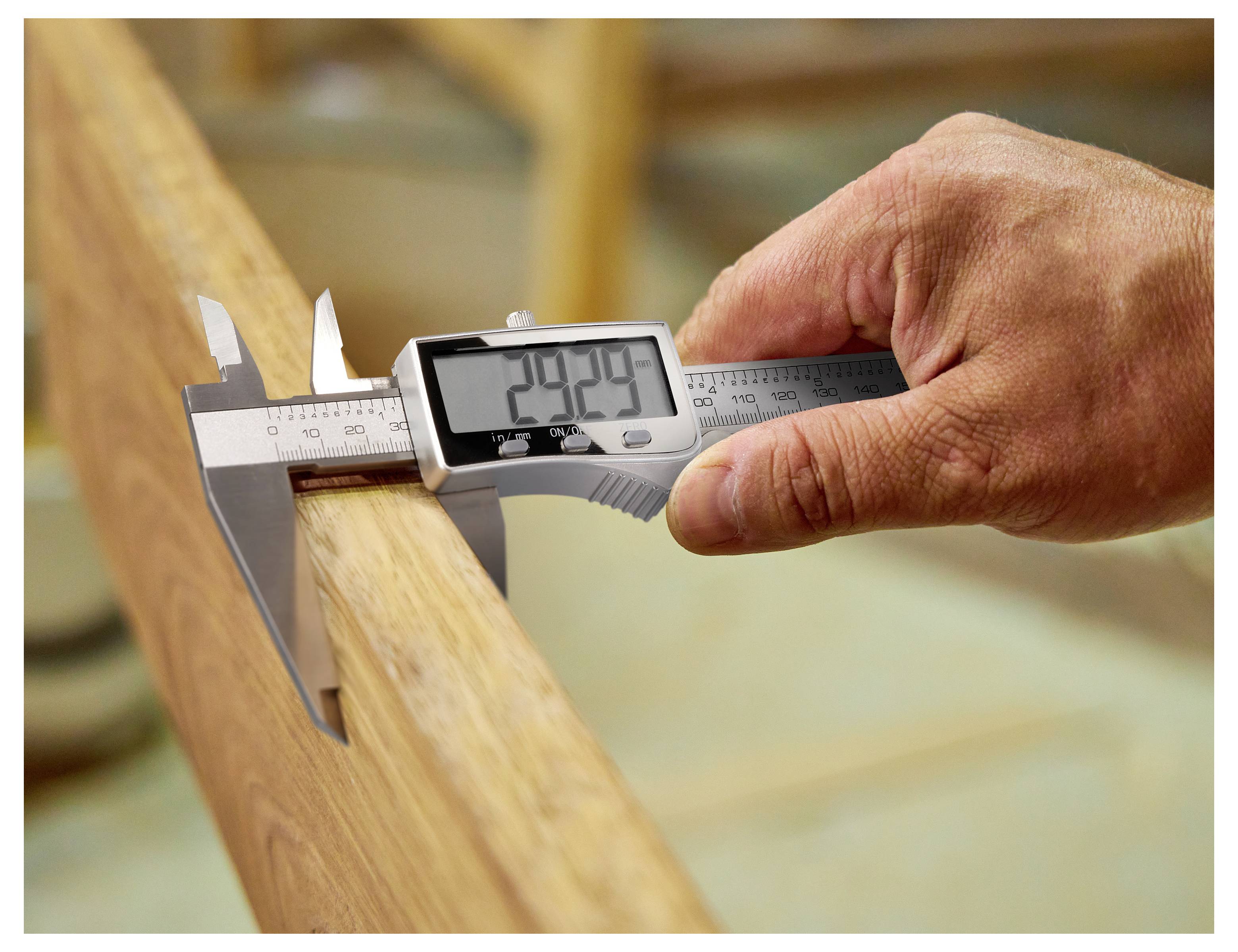 Eine Hand verwendet eine digitale Schieblehre, um die Dicke eines Holzbretts zu messen, wobei auf dem Bildschirm „29,94