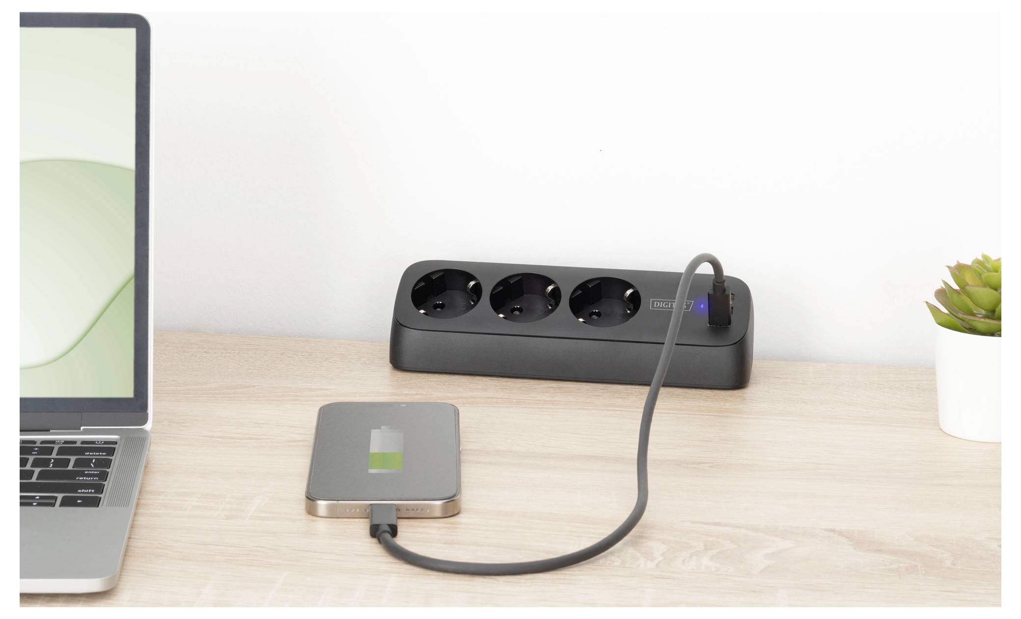 Ein Laptop auf einem Schreibtisch ist durch ein Kabel mit einem externen Gerät verbunden, das ein Ladesymbol anzeigt. Eine Steckerleiste mit drei Anschlüssen befindet sich im Hintergrund.