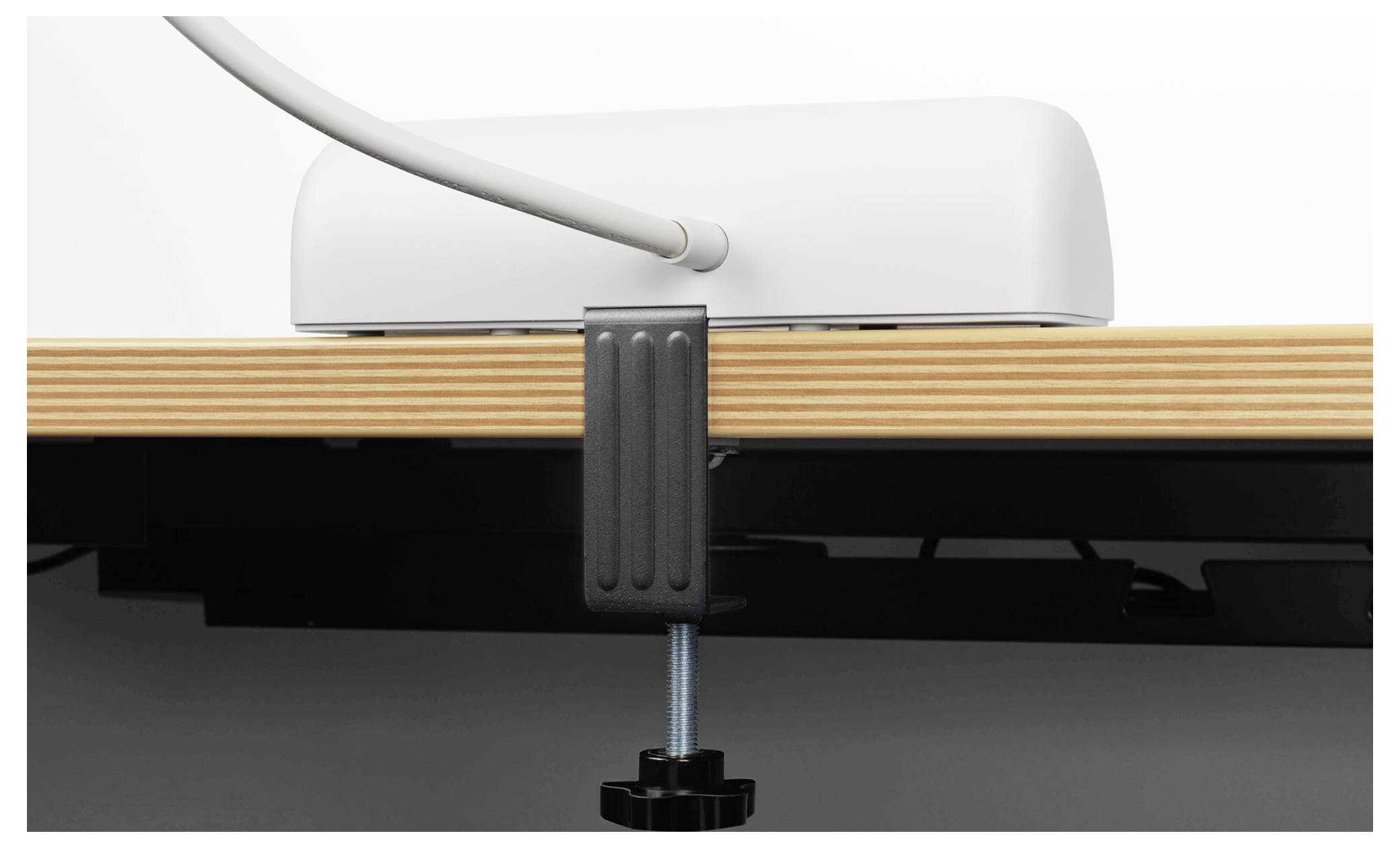 Ein weißes Gerät steht auf einem Holzschreibtisch und ist mit einer schwarzen Klammer gesichert. Ein Kabel ragt aus dem Gerät heraus, was darauf hindeutet, dass es angeschlossen oder mit Strom versorgt ist.