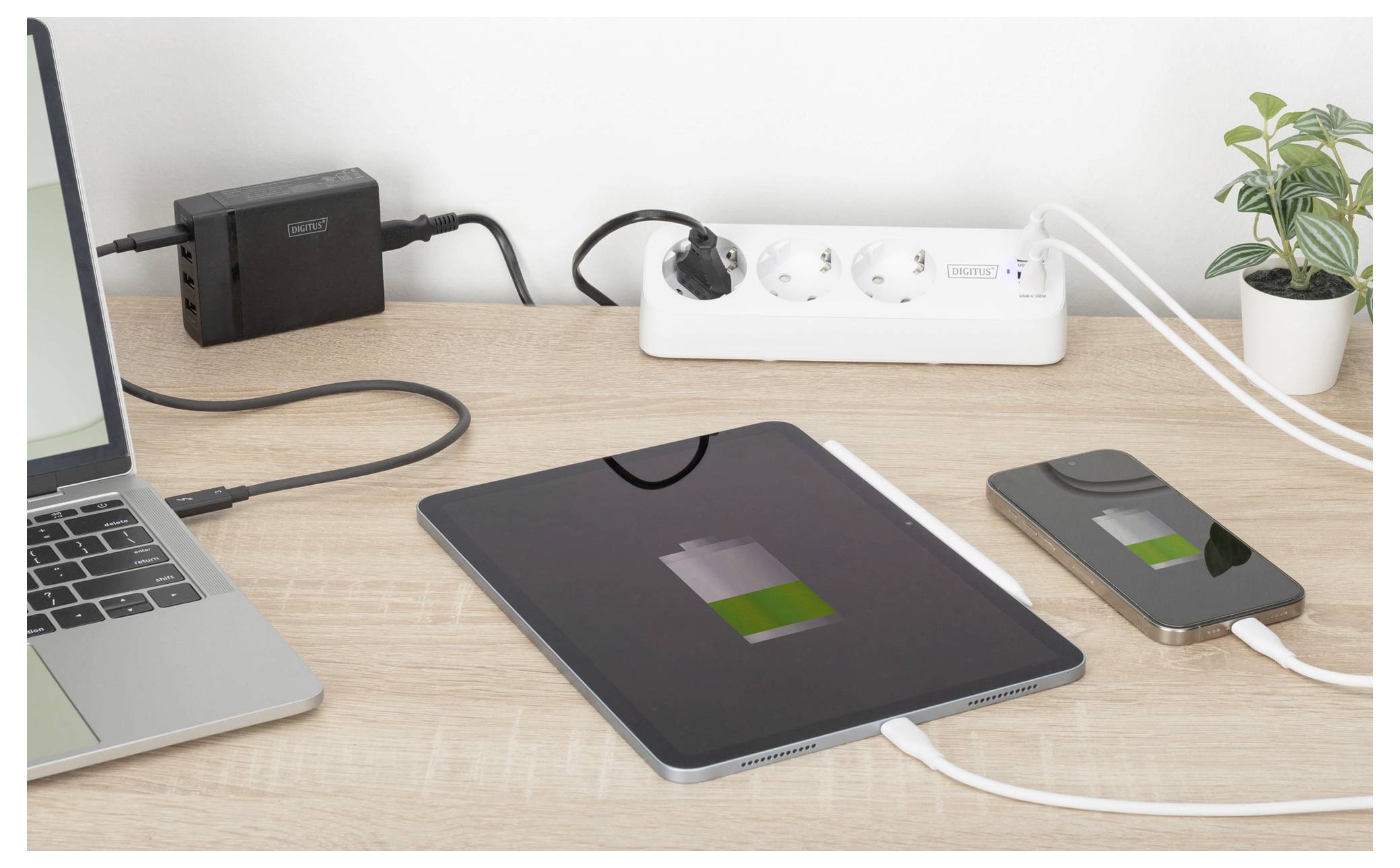 Ein Schreibtisch zeigt ein Ladetablett und ein Smartphone, die an eine Steckdosenleiste angeschlossen sind, mit einer Pflanze und einem Laptop in der Nähe, was auf das Laden von Geräten hinweist.