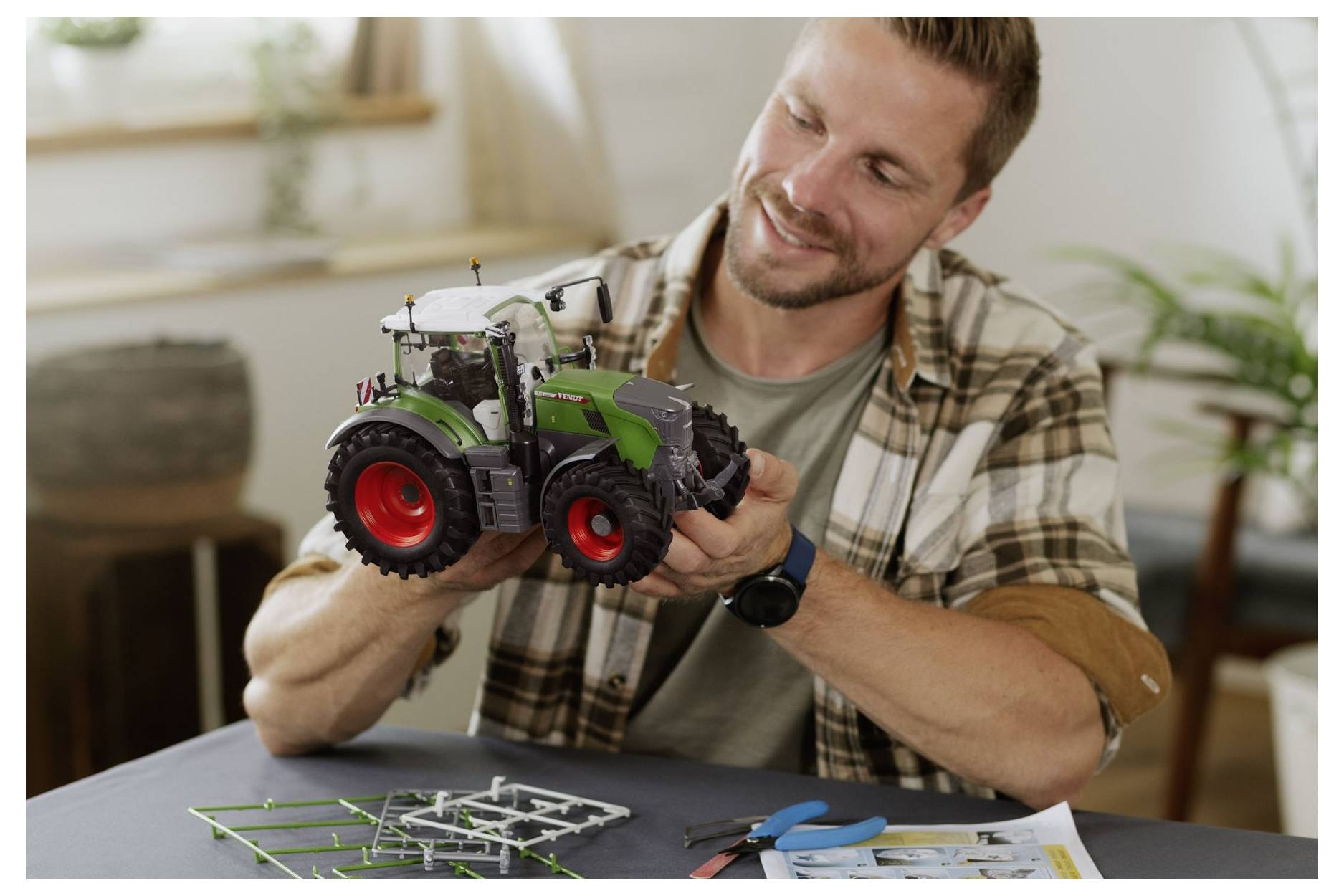 Ein Mann montiert einen Modell-Traktor und hält ihn in seinen Händen, während er an einem Tisch mit Werkzeugen und Montageanleitung sitzt.