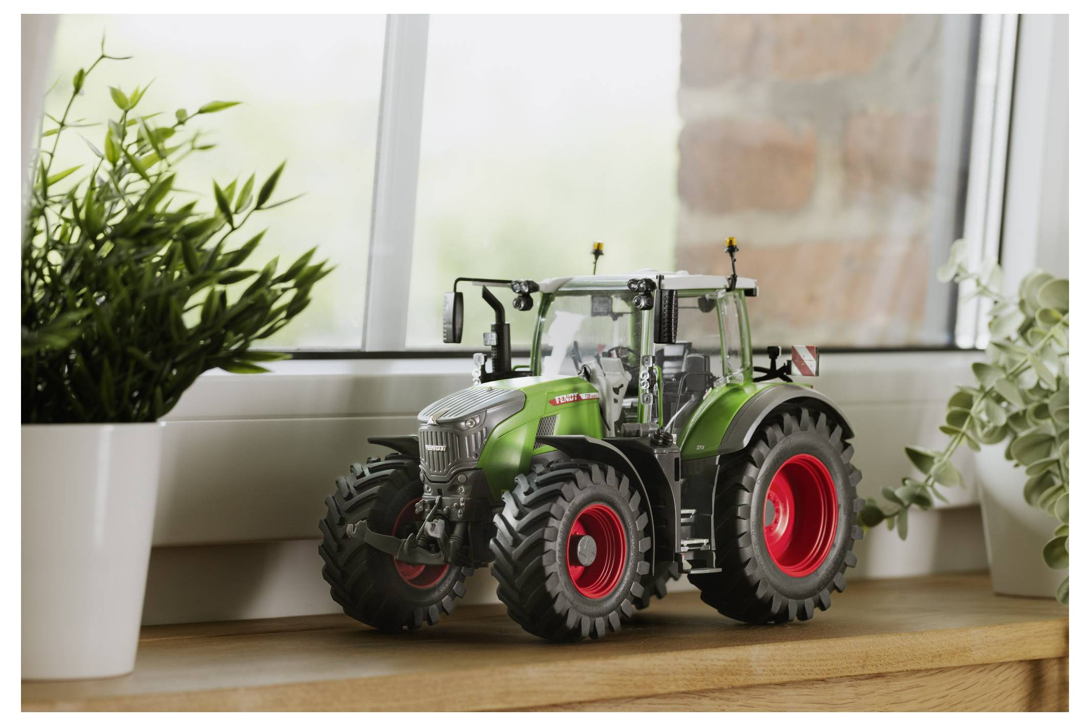 Ein Modell-Traktor mit großen roten Rädern steht auf einem Holzregal neben einem Fenster, umgeben von grünen Pflanzen.