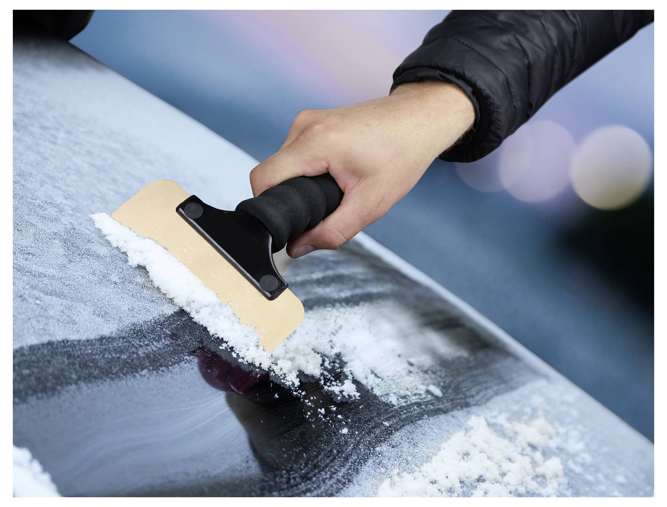 Eine Hand, die einen Eiskratzer hält, entfernt Reif von einer Autoscheibe und bahnt sich einen Weg durch das Eis, was Wartung bei kaltem Wetter signalisiert.