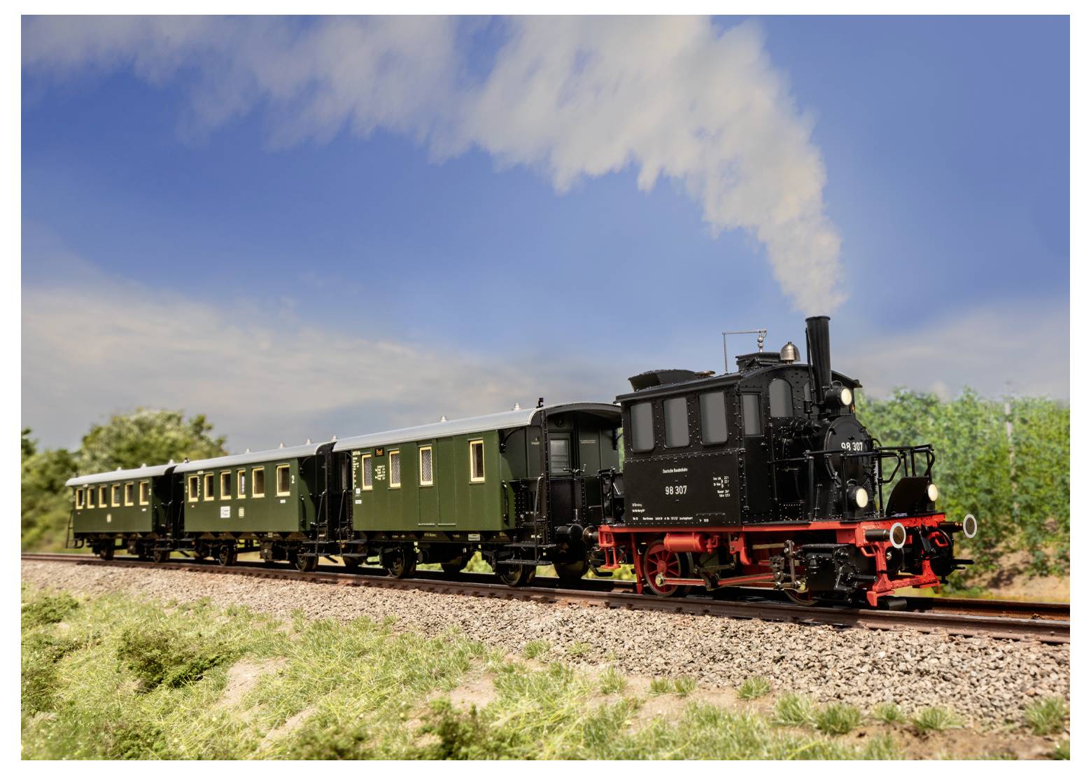 Eine kleine Dampflokomotive zieht zwei grüne Personenwagen auf einer ländlichen Bahnstrecke unter einem klaren blauen Himmel mit einigen Wolken.