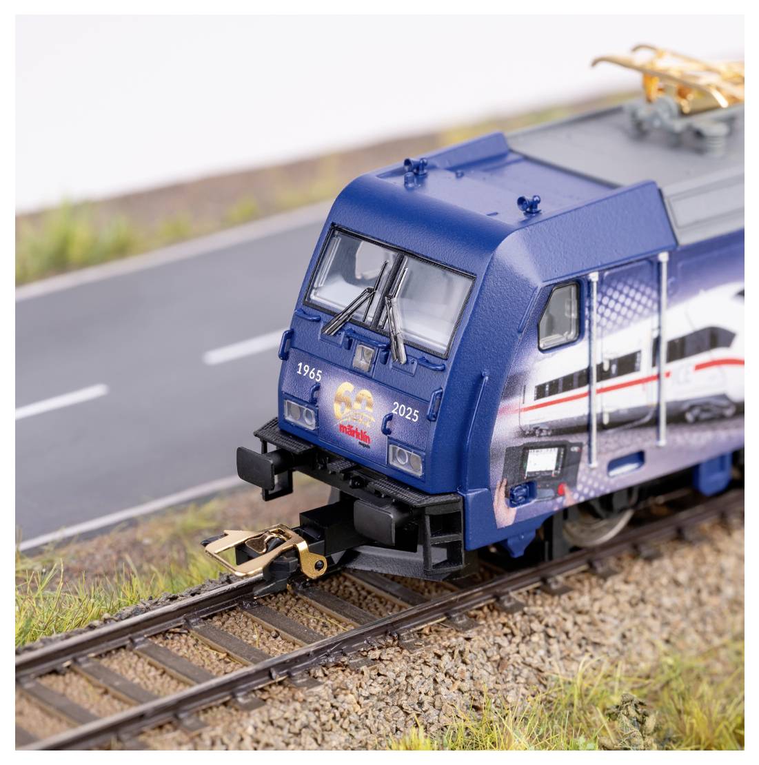 Modelleisenbahn auf Schienen neben einer Straße, mit blauer Lokomotive, die die Markierung '1965-2025' trägt und 60 Jahre eines Eisenbahnunternehmens feiert.