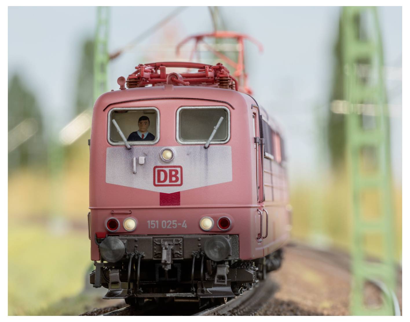 Ein roter Zug mit dem 'DB'-Logo und der Nummer '151 025-4' an der Front fährt auf Schienen, mit einem unscharfen Hintergrund von Grünflächen.