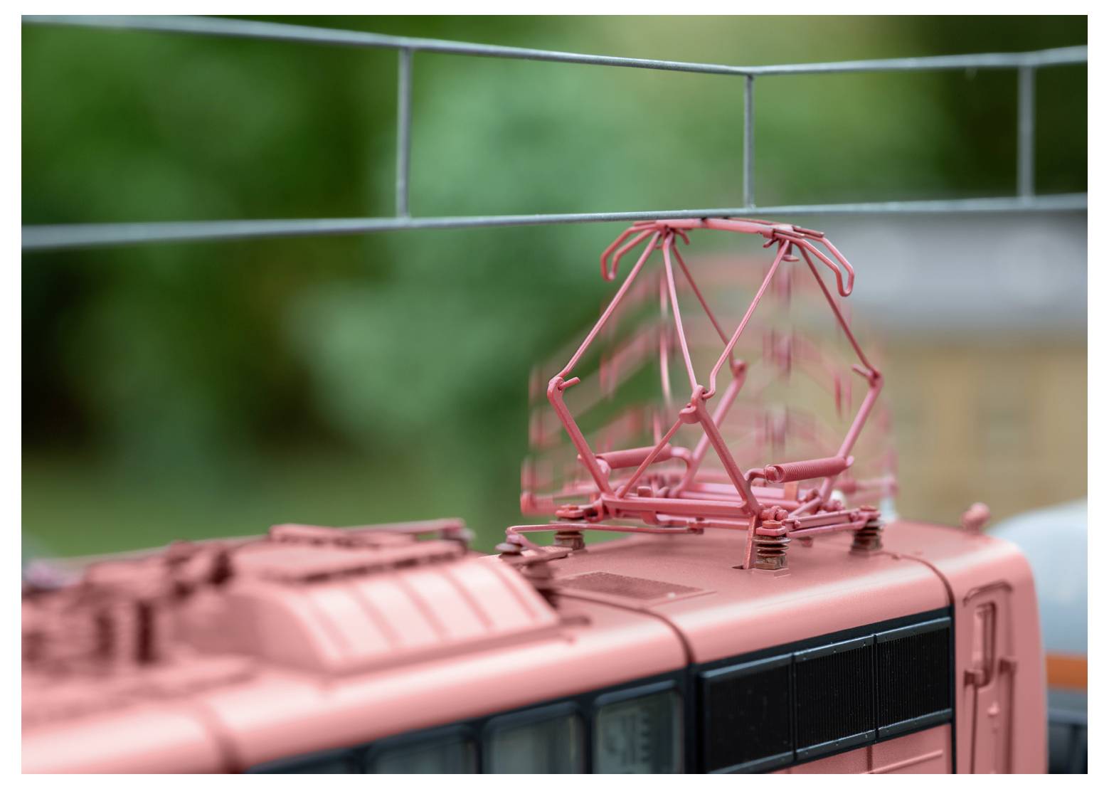 Eine Nahaufnahme des Daches eines Modellzugs mit einem roten Stromabnehmer vor einem unscharfen grünen Hintergrund.