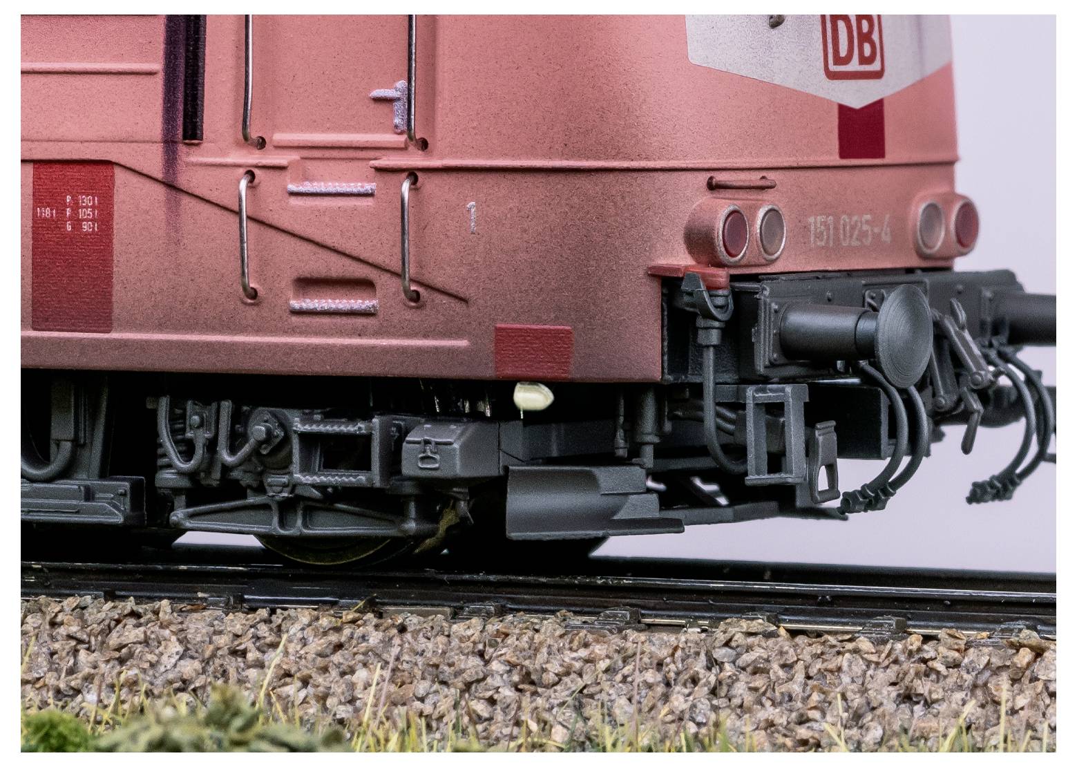 Nahaufnahme des hinteren Teils einer roten Zuglokomotive, die detaillierte mechanische Komponenten und das 'DB'-Logo zeigt. Die Gleise sind unter der Lokomotive sichtbar.